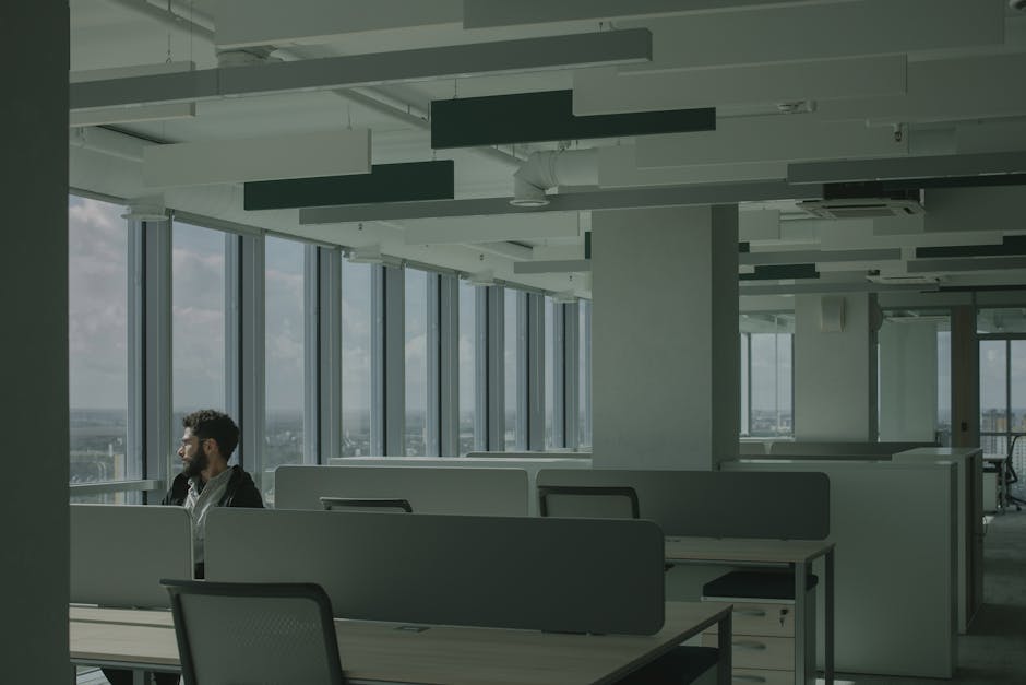 A man sits alone in a modern office space with large windows overlooking a cityscape